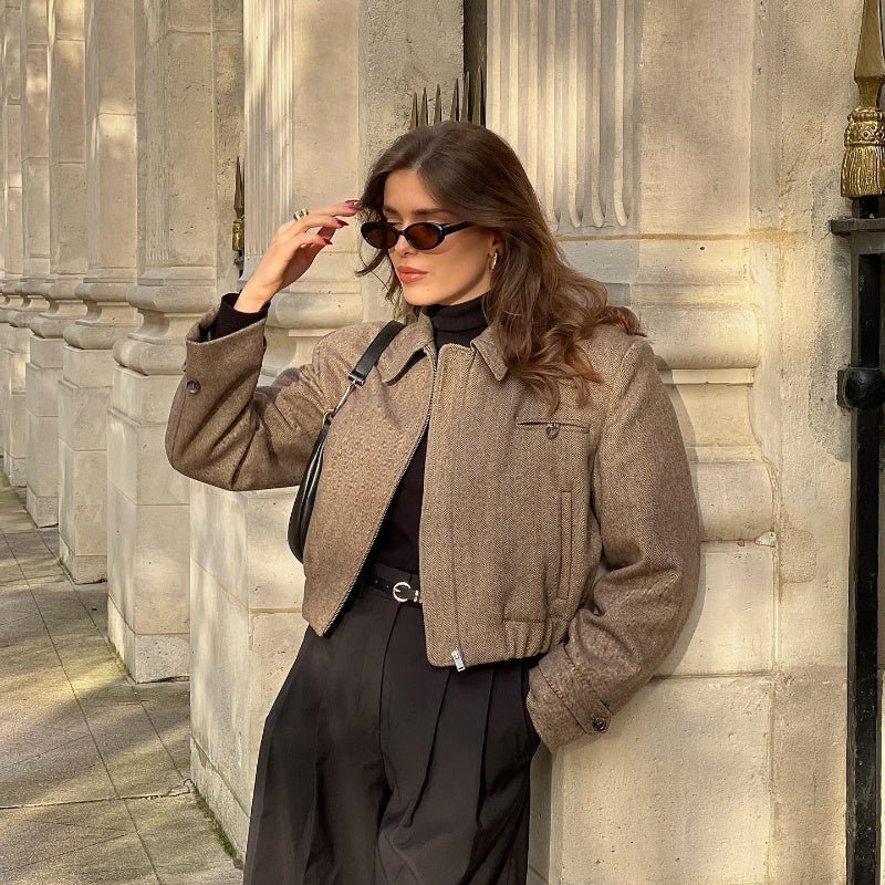 A woman wearing a coat and sunglasses leans casually against a wall, exuding a relaxed and stylish demeanor.